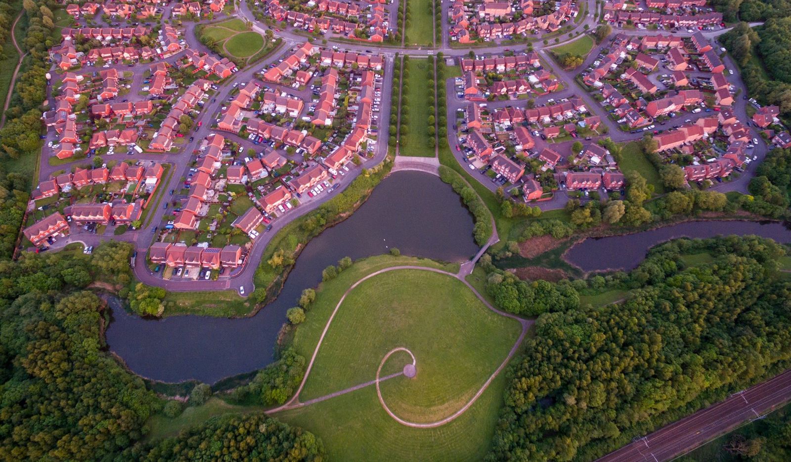 An aerial photograph at golden hour showing the New Bold estate above a wooded river, a lake, an open meadow and a spiral mound earthwork — the registered open space at the heart of this petition.